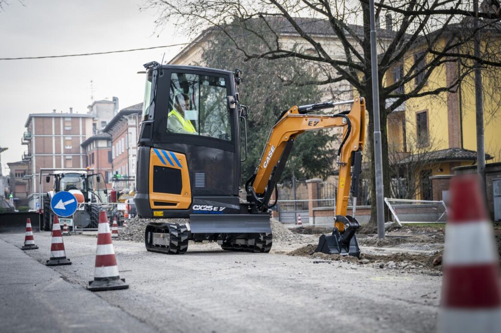 Minikoparka CASE CX25 EV podczas prac drogowych w mieście – operator wykonuje wykop w pasie drogowym między pachołkami, w otoczeniu budynków i infrastruktury miejskiej.